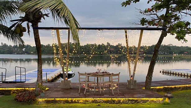 Terraza junto al lago con una mesa y sillas, rodeada de luces colgantes y palmeras.