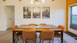 Een eethoek met een houten tafel, oranje stoelen en een kroonluchter. Boven de tafel hangen drie ingekaderde foto's aan de muur.