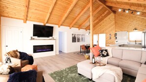 Een ruime woonkamer met een hoog houten plafond, een flatscreen televisie boven een open haard en een moderne keuken op de achtergrond.