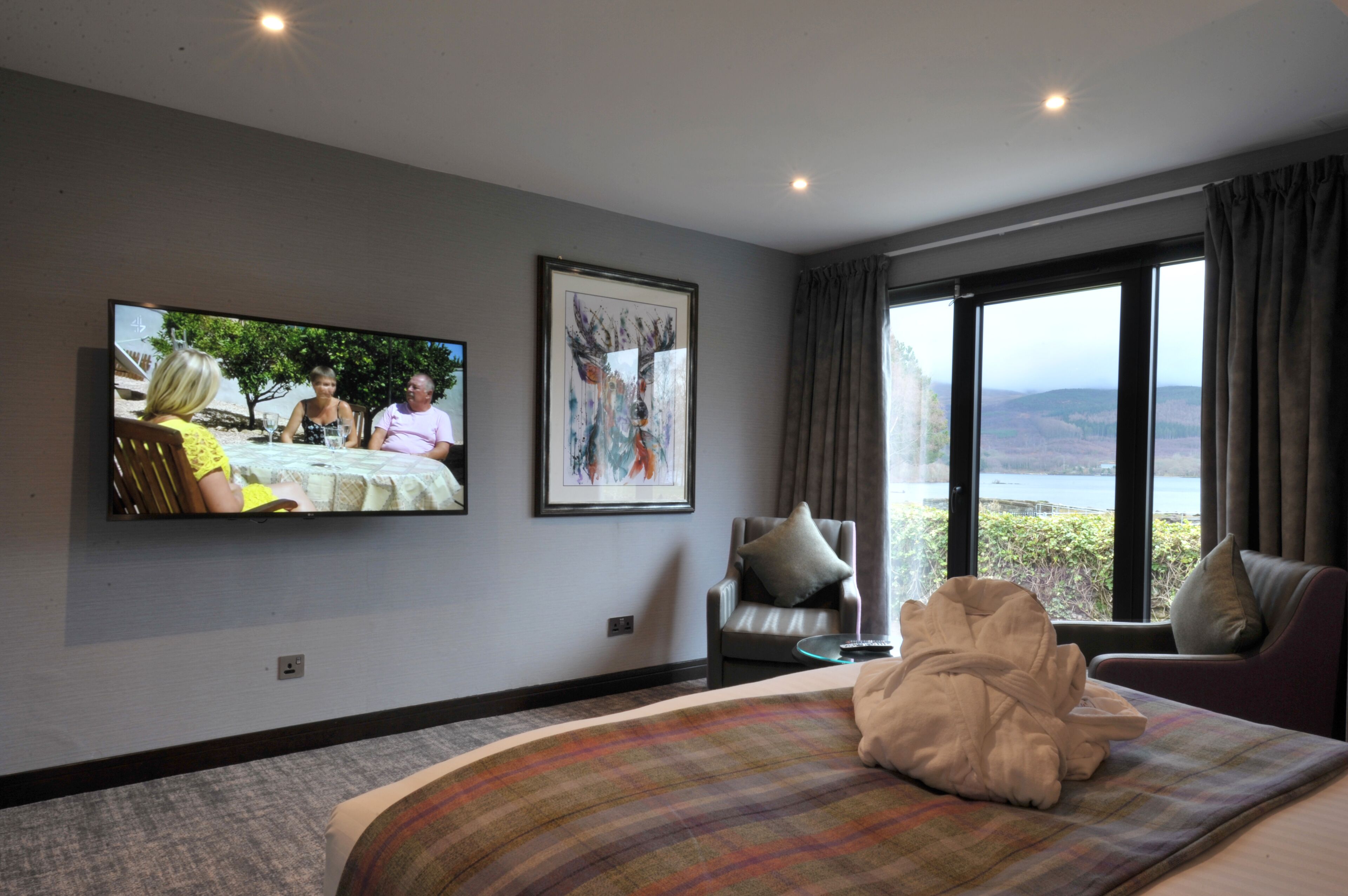 Una moderna camera d'hotel con un letto grande, una televisione fissata al muro, un quadro astratto incorniciato e vista su un lago attraverso ampie finestre.