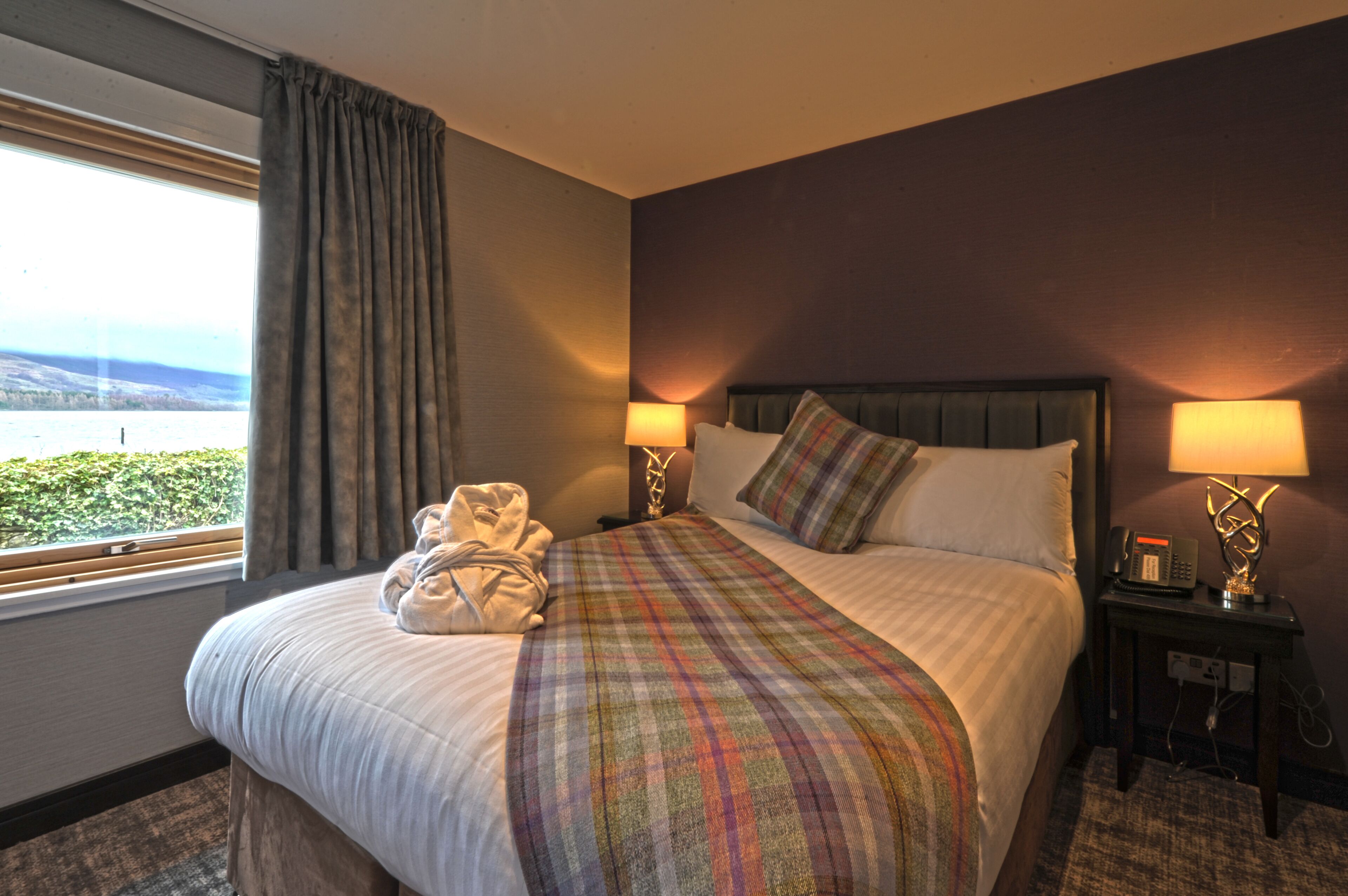 A hotel room with a large bed, two bedside lamps, a telephone, and a view of a body of water through a window with curtains.