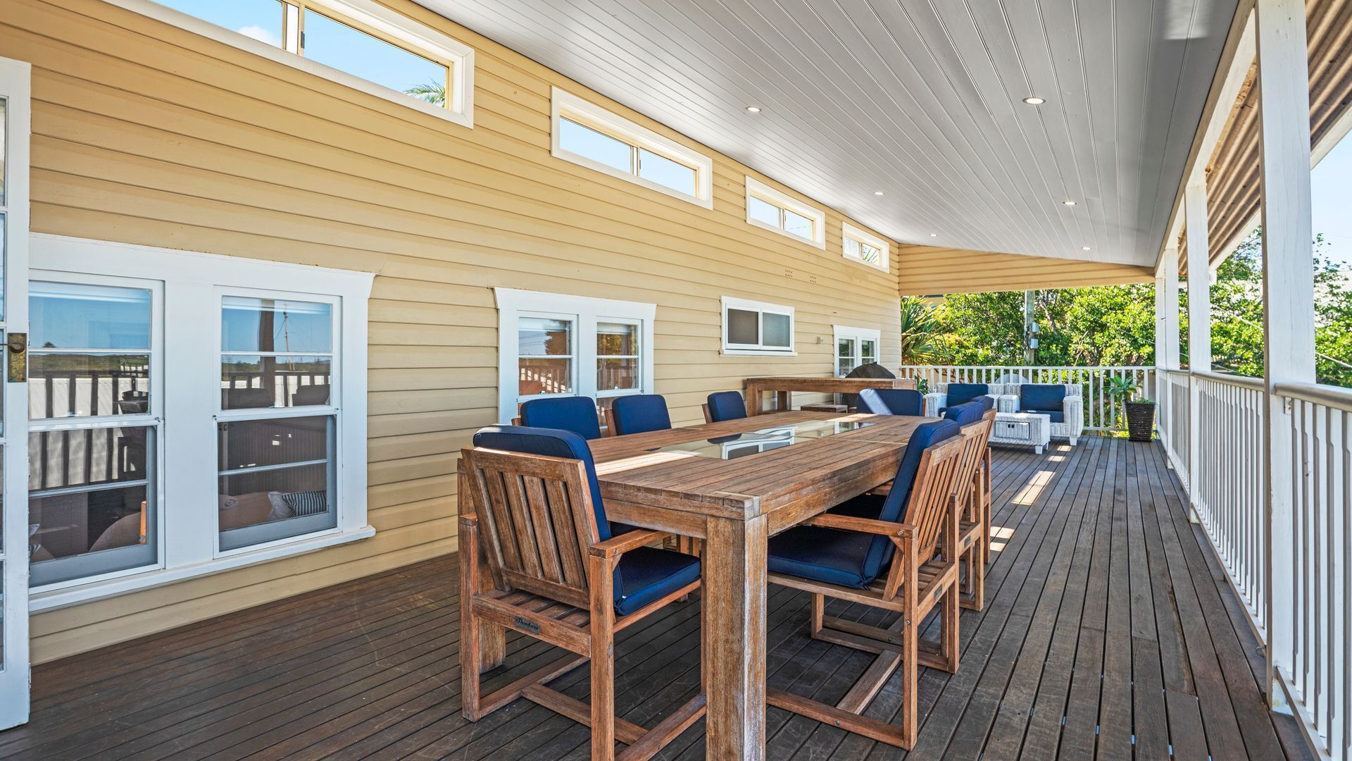 Um deck externo coberto com mesa e cadeiras de madeira, guarda-corpos brancos e várias janelas.