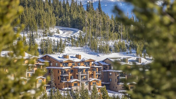 A modern lodge with multiple wooden buildings, surrounded by snow-covered terrain and dense evergreen trees. - Penthouse | Close to Shuttle | Hot Tub (Big Sky)
