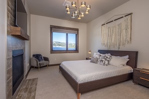 A bedroom with a large bed, a fireplace, a window with a view, and a chandelier. - Spend Summer in Big Sky! Condo with Lone Peak Views (Big Sky)