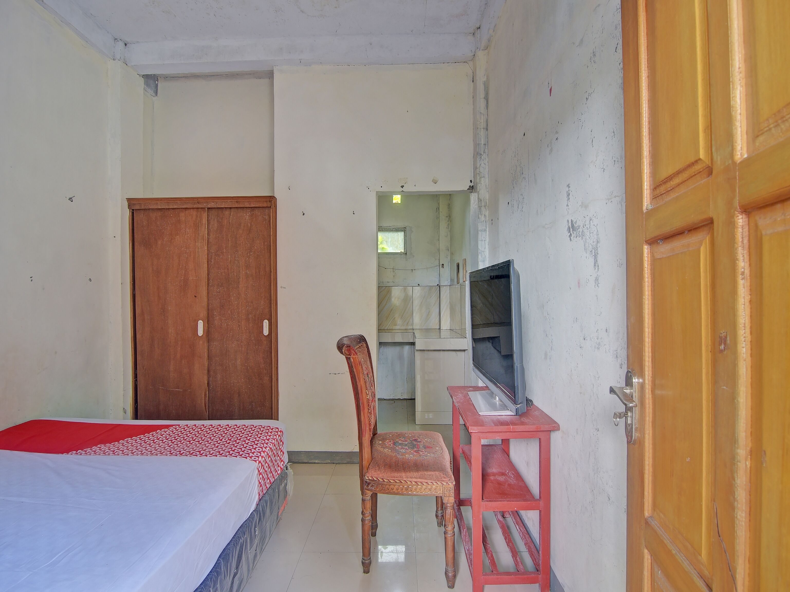 Quarto com uma cama, guarda-roupa de madeira, televisão sobre um suporte vermelho e uma cadeira.