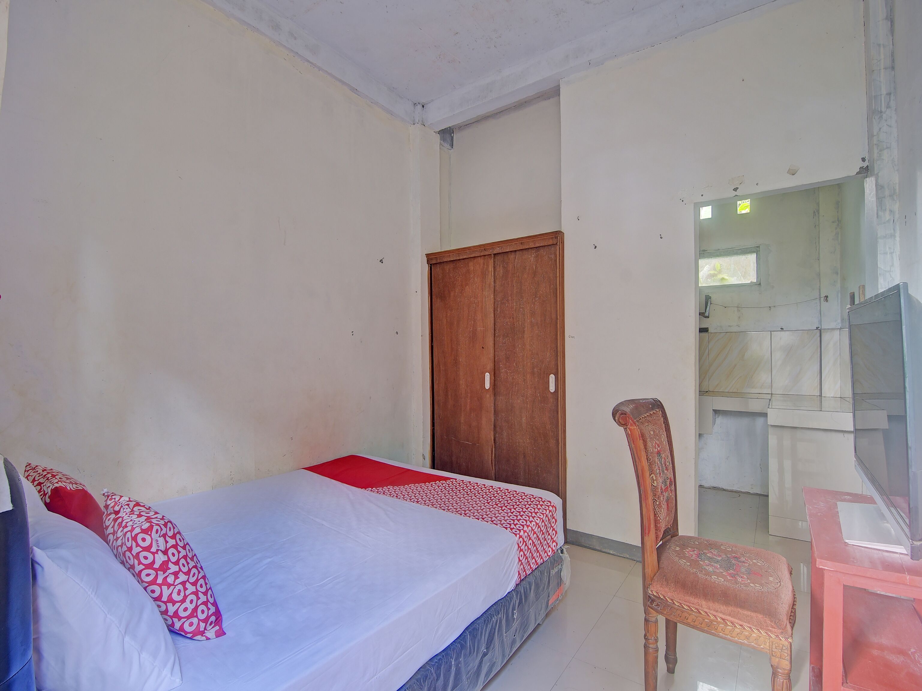 A bedroom with a bed, a wooden wardrobe, a chair, and a television on a stand.