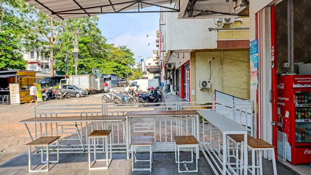 An outdoor seating area with white metal tables and benches, a red Coca-Cola vending machine, and a street view with parked vehicles and buildings in the background. - Hotel O Zazezo Homestay Syariah (Tanjung Pinang)