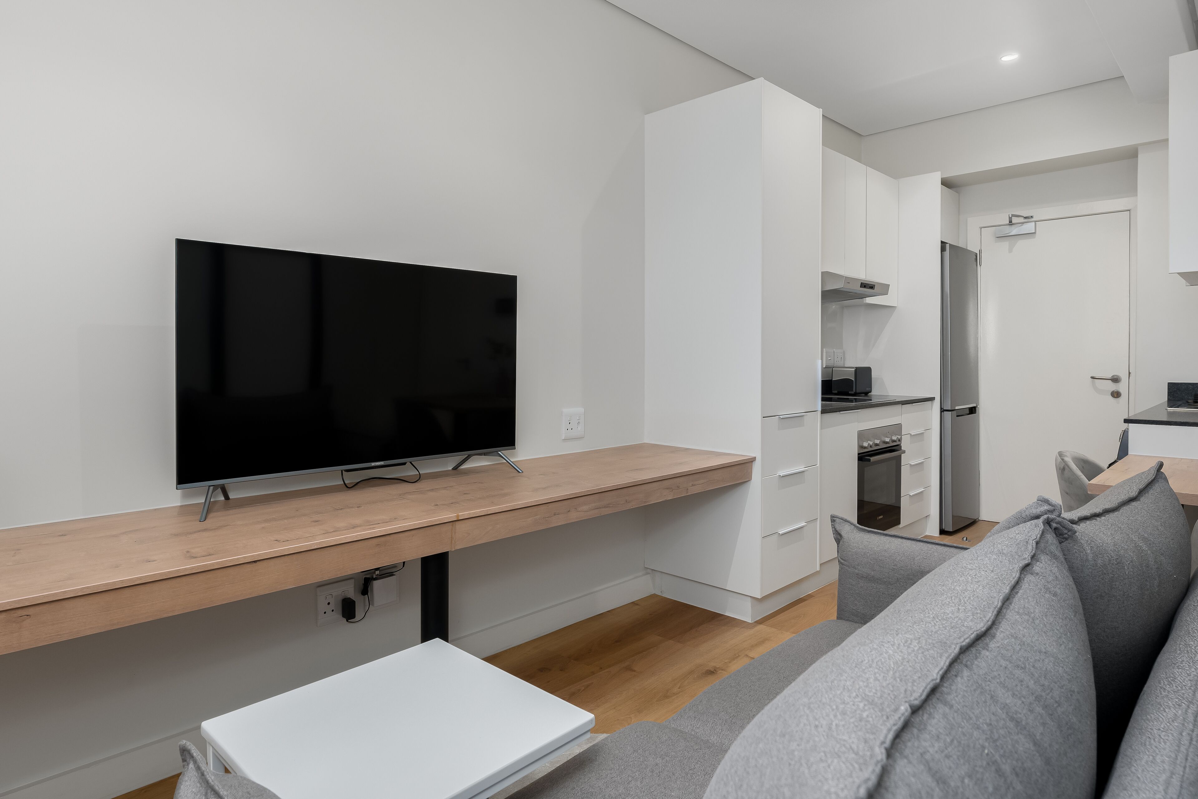 A modern living room with a flat-screen TV, a wooden desk, a grey sofa, and a kitchen area in the background.
