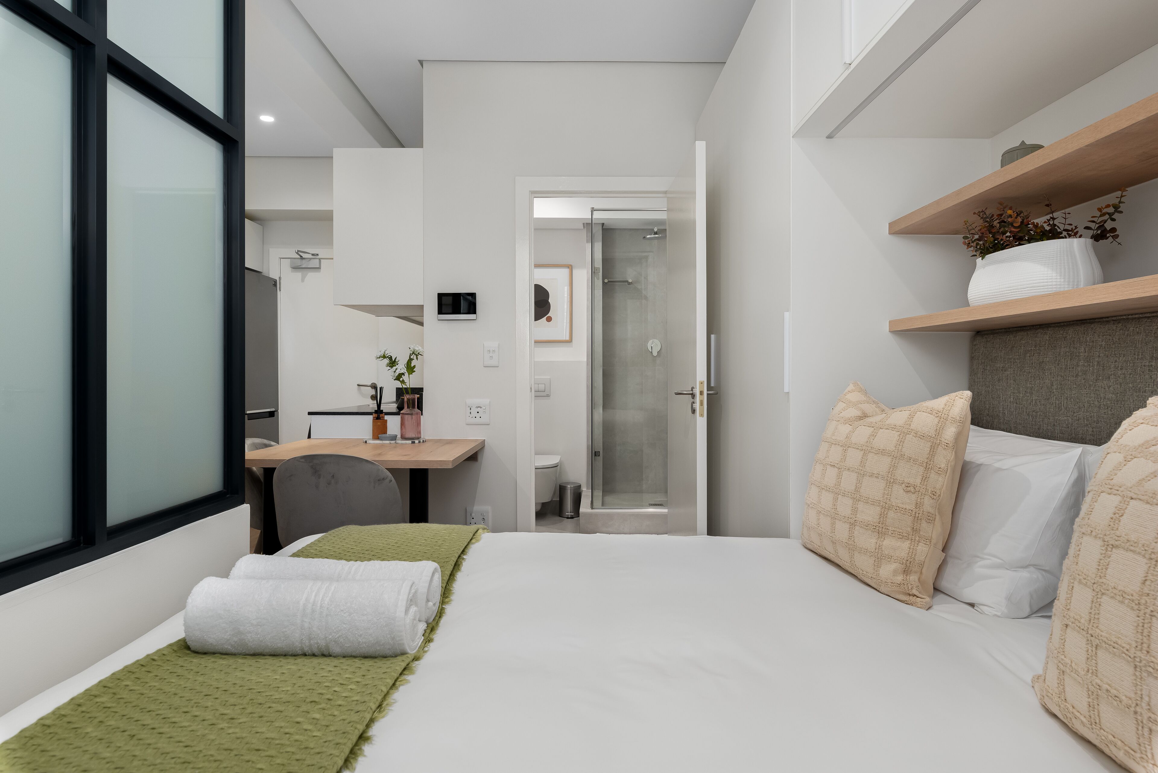 A modern bedroom with a bed, green throw, white pillows, a desk, and a shower area.