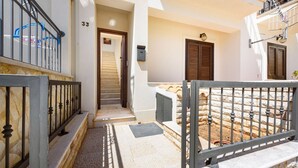 A narrow outdoor corridor with a stone floor, a metal gate, and a door leading to a staircase.