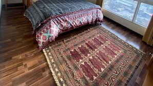 A bedroom with a large bed, a ceiling fan, a nightstand, a window with a view, and a patterned rug. - Luxurious Apartment with Fascinating Margalla View (Islamabad)