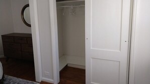 Ein Zimmer mit einer geschlossenen weißen Tür, einem hölzernen Schminktisch und einem Kleiderschrank mit Kleidung.