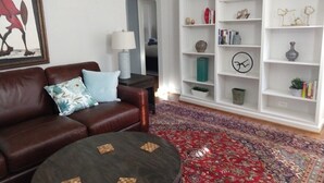 Ein Wohnzimmer mit einer braunen Ledersofa, einem runden Couchtisch und einem gemusterten Teppich. Eine weiße Bücherwand beherbergt verschiedene Dekorationselemente und ein gerahmtes Bild an der Wand.