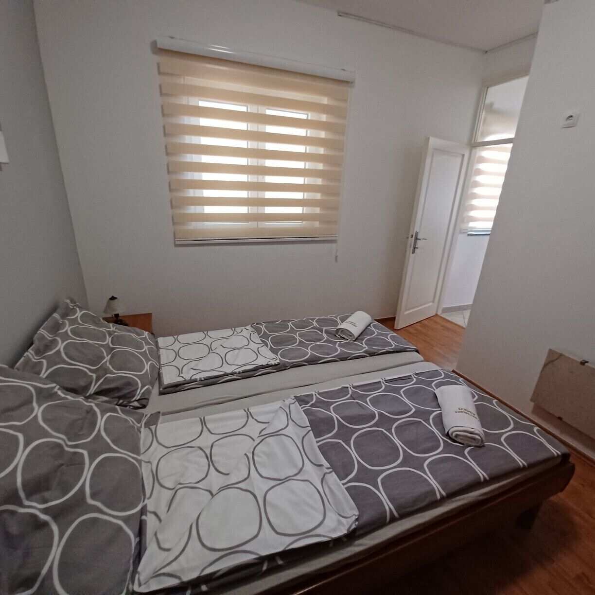 A bedroom with two beds, a window with blinds, a door, and a wooden floor.