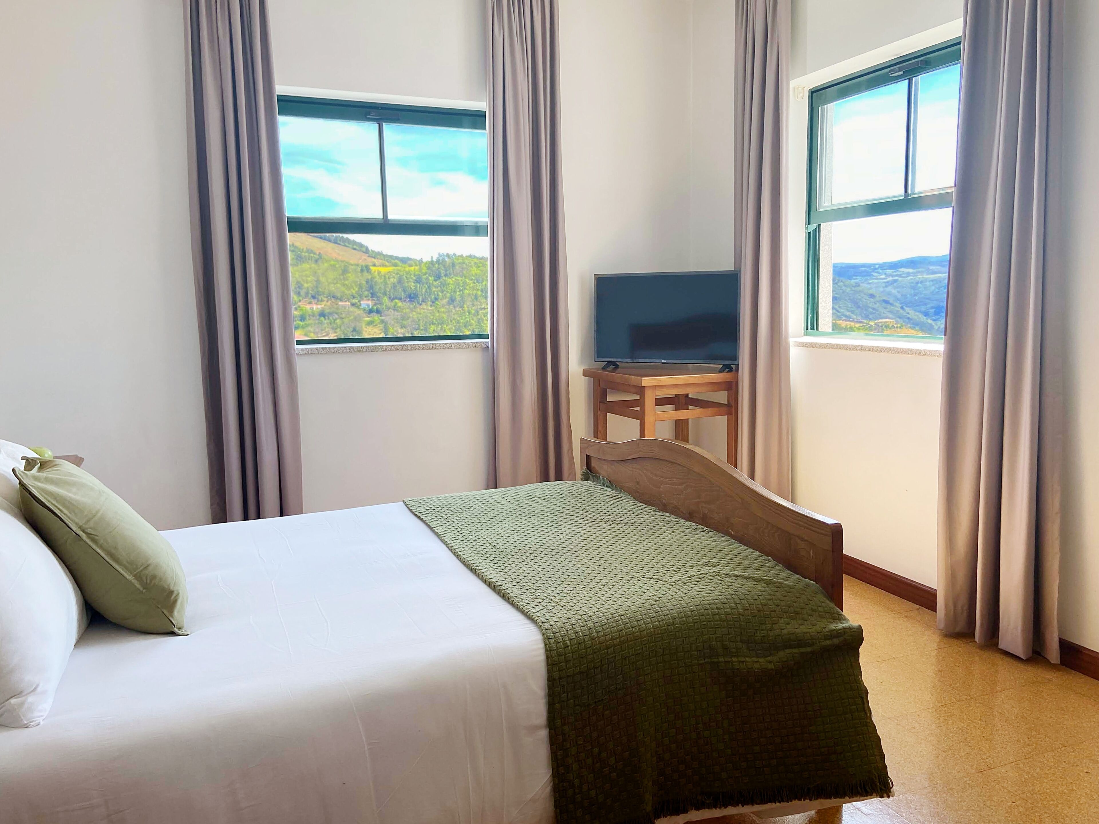 A hotel room with a bed, a TV on a small table, and large windows with curtains.
