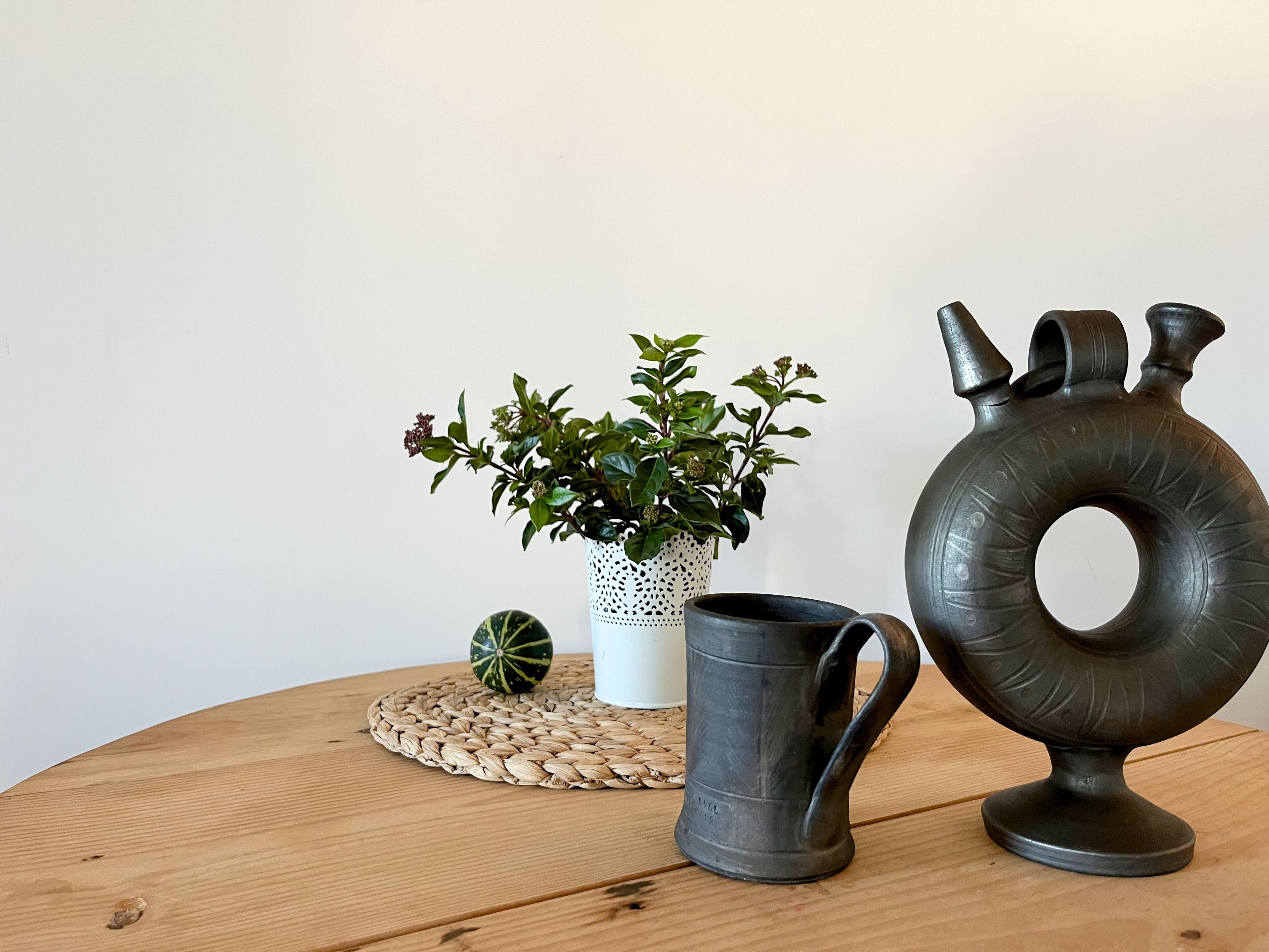 Una mesa redonda de madera con un mantel tejido, una planta en maceta, una jarra de metal y un florero de metal.