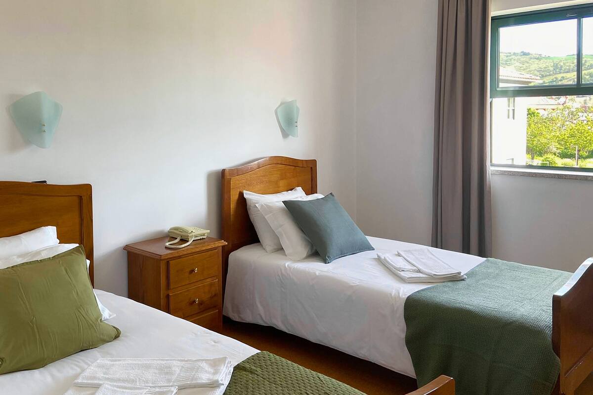 A hotel room with two beds, a wooden nightstand, a telephone, and a window with a view of greenery.