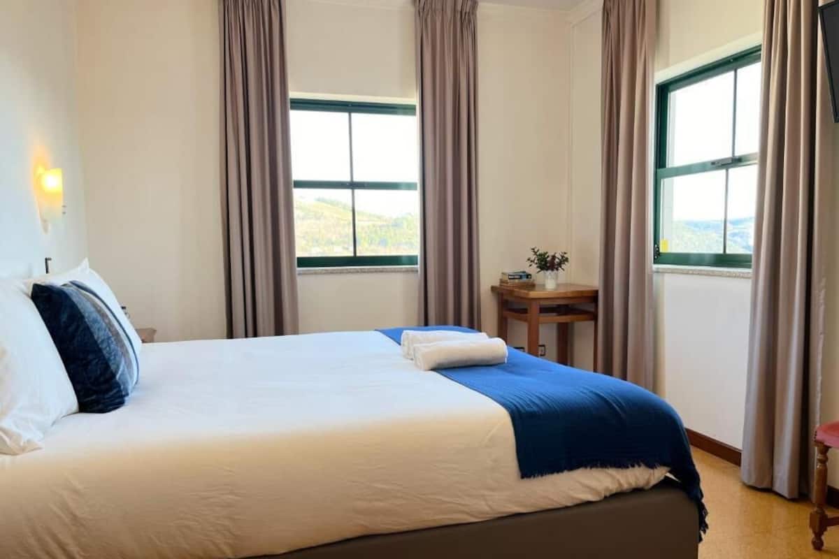A neatly made bed with white and blue bedding, a small wooden table with a vase, and a view through a window with curtains.