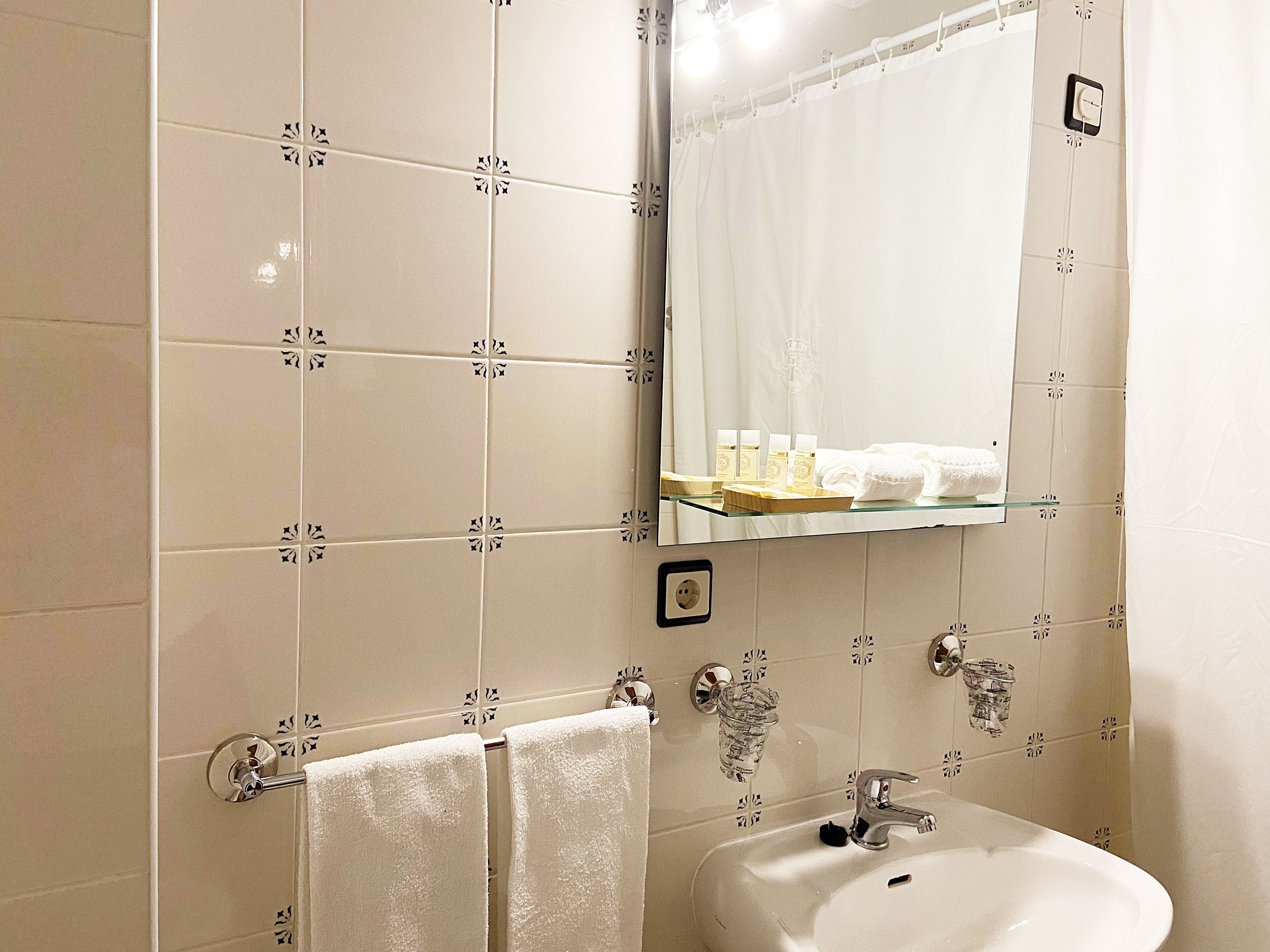 A bathroom with white tiled walls, a glass shelf with toiletries, a sink, and two towels hanging on a rack.