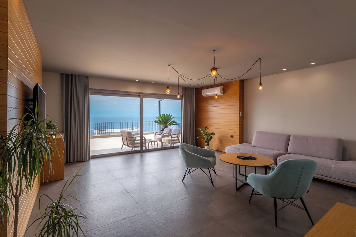 A modern living room with a sofa, two armchairs, a coffee table, and a TV. There is a balcony with a view of the sea.