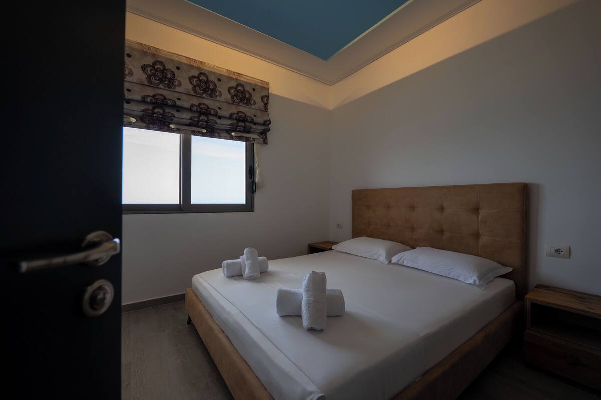 A hotel room with a large bed, two pillows, a wooden headboard, and a window with a decorative valance.