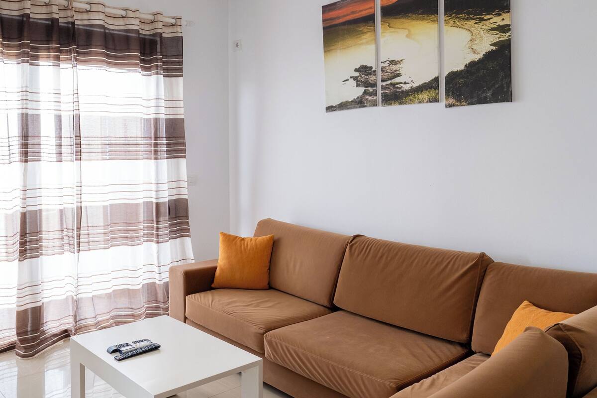 A modern living room with a brown sofa, a white coffee table, and a hanging lamp. There are three framed pictures on the wall.