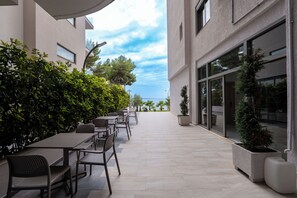An outdoor patio with tables and chairs, a hedge, and a building with large windows. - Franklin Rooms (Sarandë)