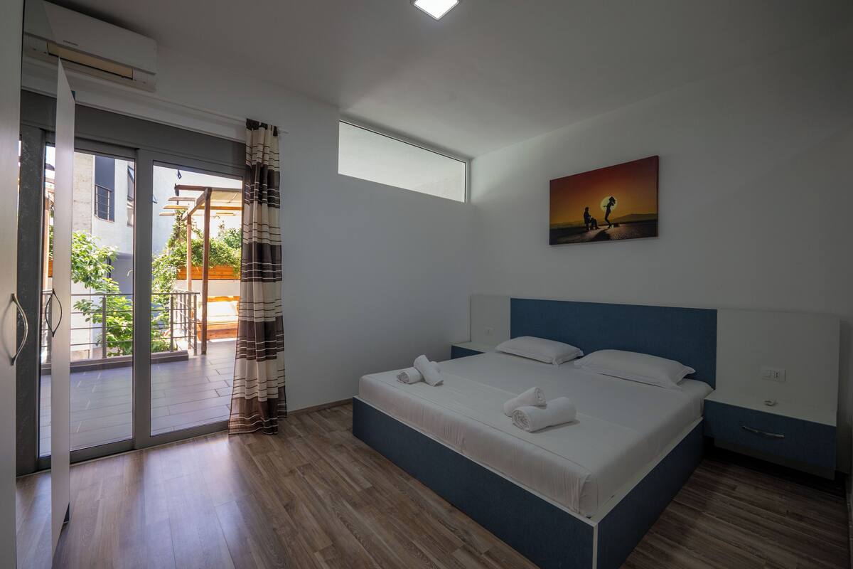 A bedroom with a bed, a window, a painting, and a view of the outside.
