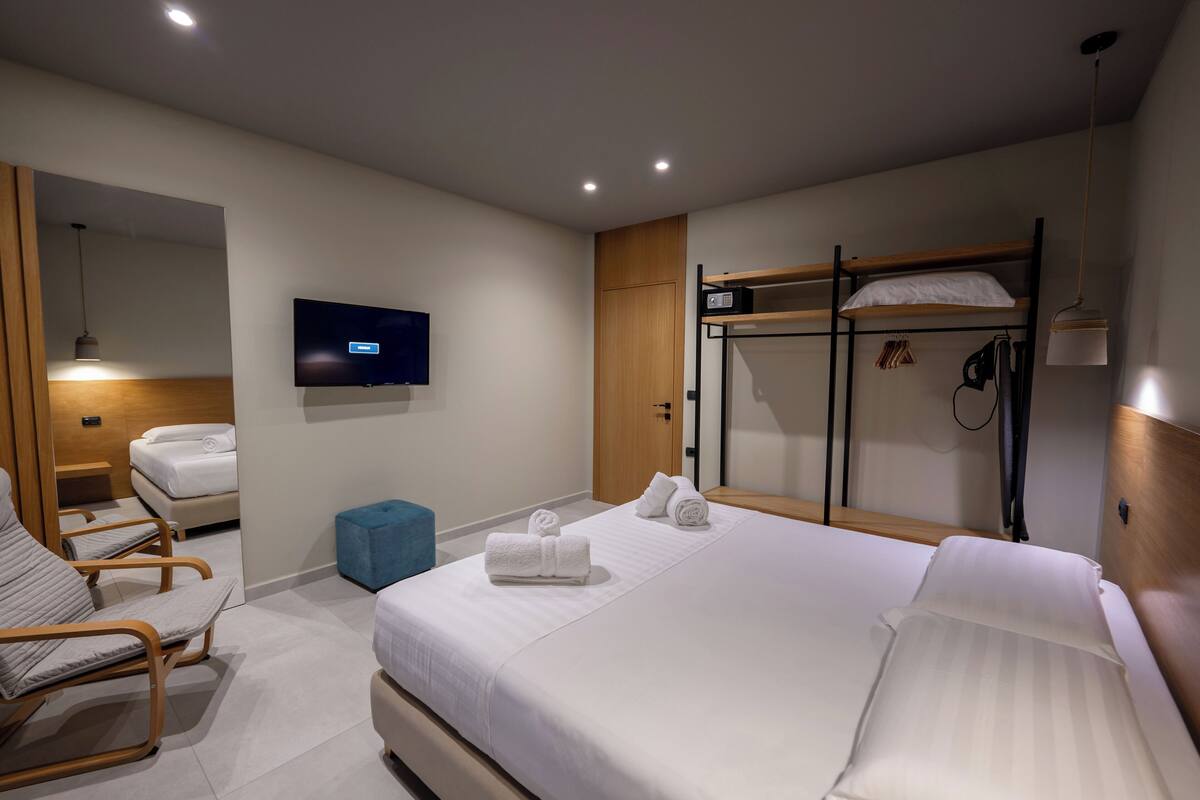 A modern hotel room with a large bed, a TV, a bunk bed, and a seating area with chairs.