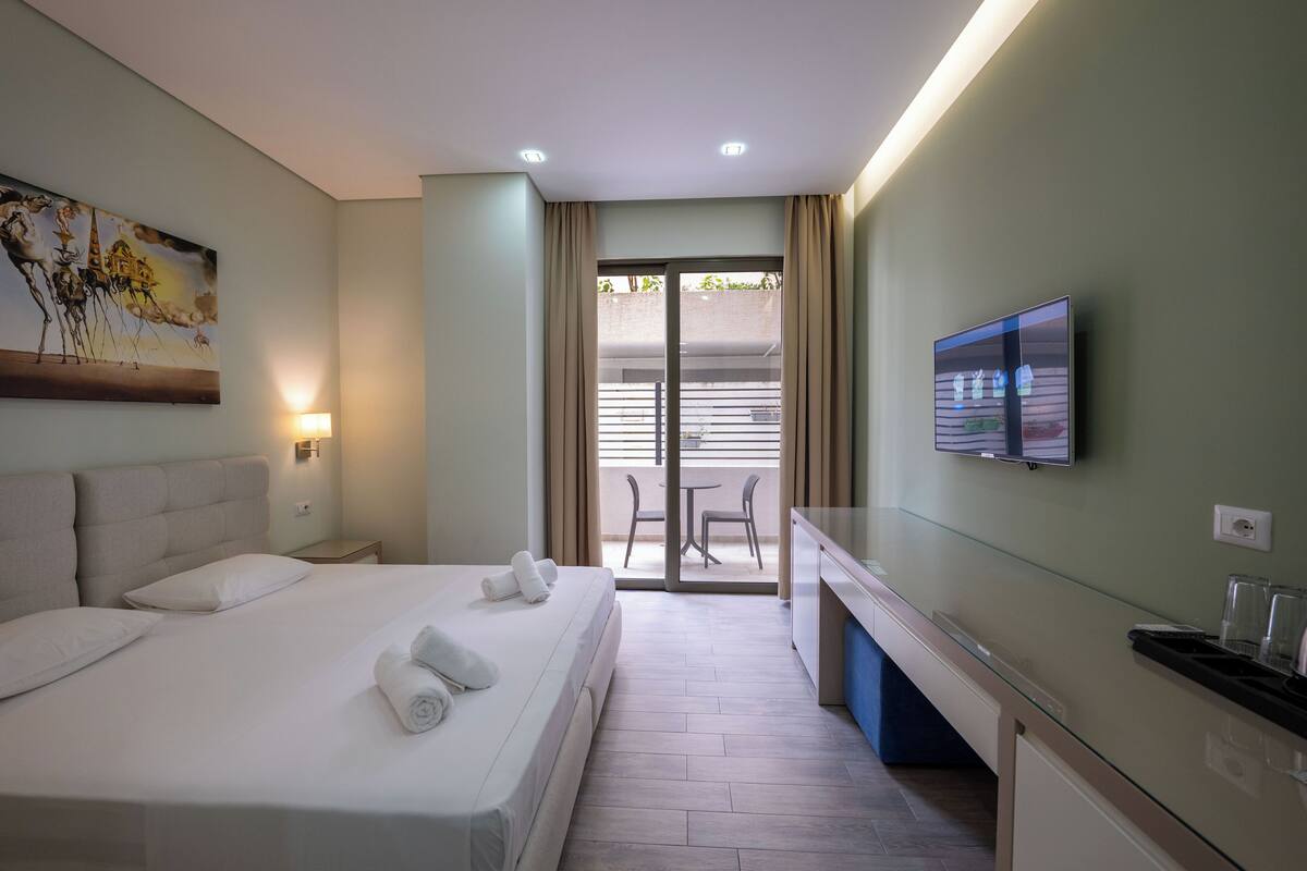 A hotel room with a bed, a desk, a television, and a balcony with chairs.