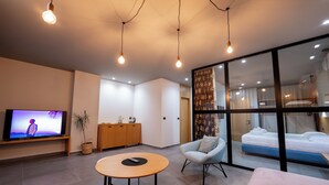 A modern hotel room with a flat-screen TV, a round wooden table, a light blue armchair, a bed, and a large window with a view of the city. - Franklin Rooms (Sarandë)