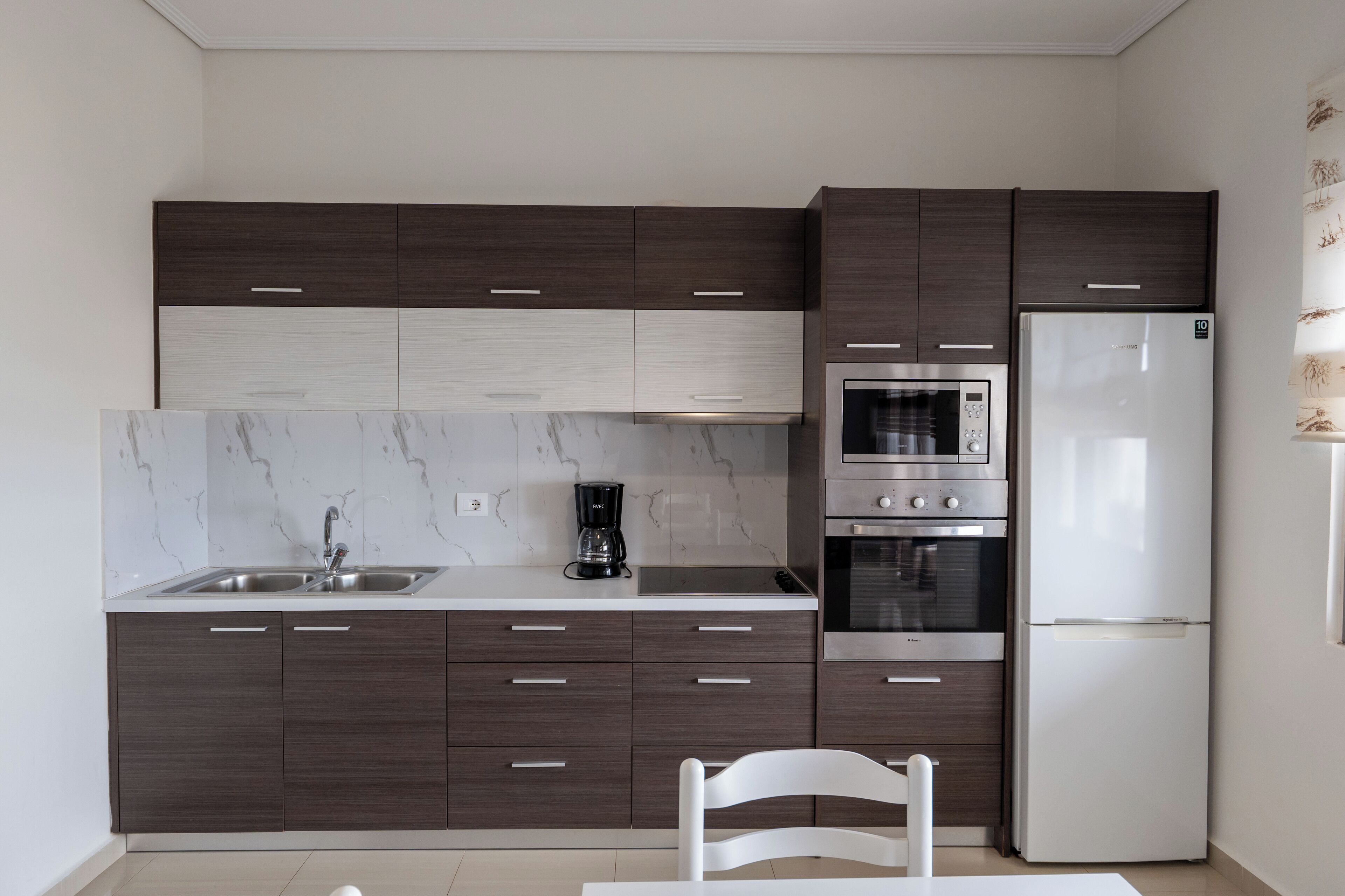 A modern kitchen with dark cabinetry, a white countertop, a stainless steel sink, a coffee maker, an oven, and a refrigerator.