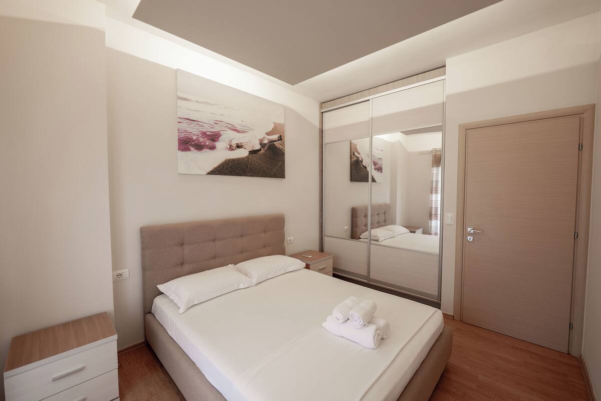 A modern bedroom with a large bed, a bedside table, a nightstand, a wardrobe, and a mirror.