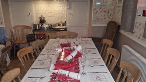 A dining room with a long table set for a meal, chairs around it, and a kitchen in the background.