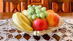 Une table à manger surmontée d’un saladier rempli de fruits, notamment des bananes, des raisins et des pommes, le tout posé sur une nappe en dentelle.
