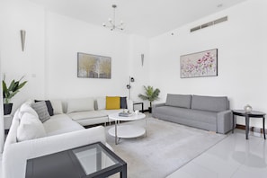 A modern living room with a grey sofa, white armchair, and a coffee table. There are two paintings on the walls and a potted plant on the floor.