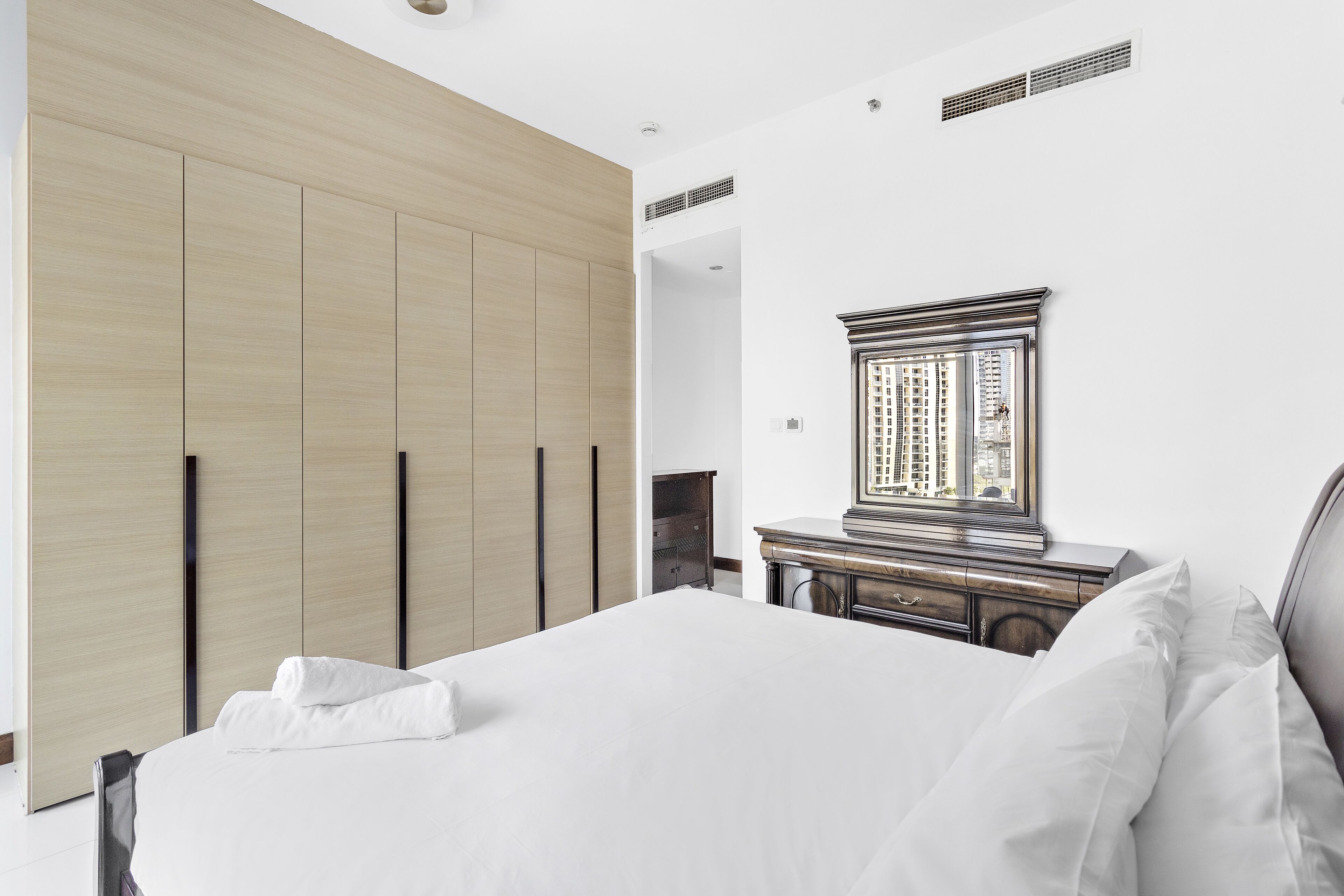 Un dormitorio con una cama grande, un armario de madera, un tocador y un espejo que refleja un paisaje urbano.