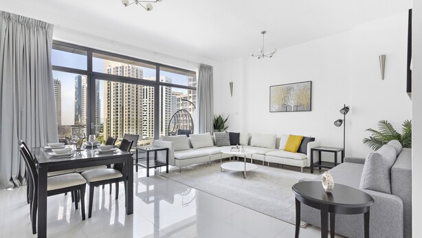 A modern living room with a dining area, a gray sofa, a coffee table, and a view of high-rise buildings. - WelHome - Urban Retreat With Terrace And City Views (Dubai)