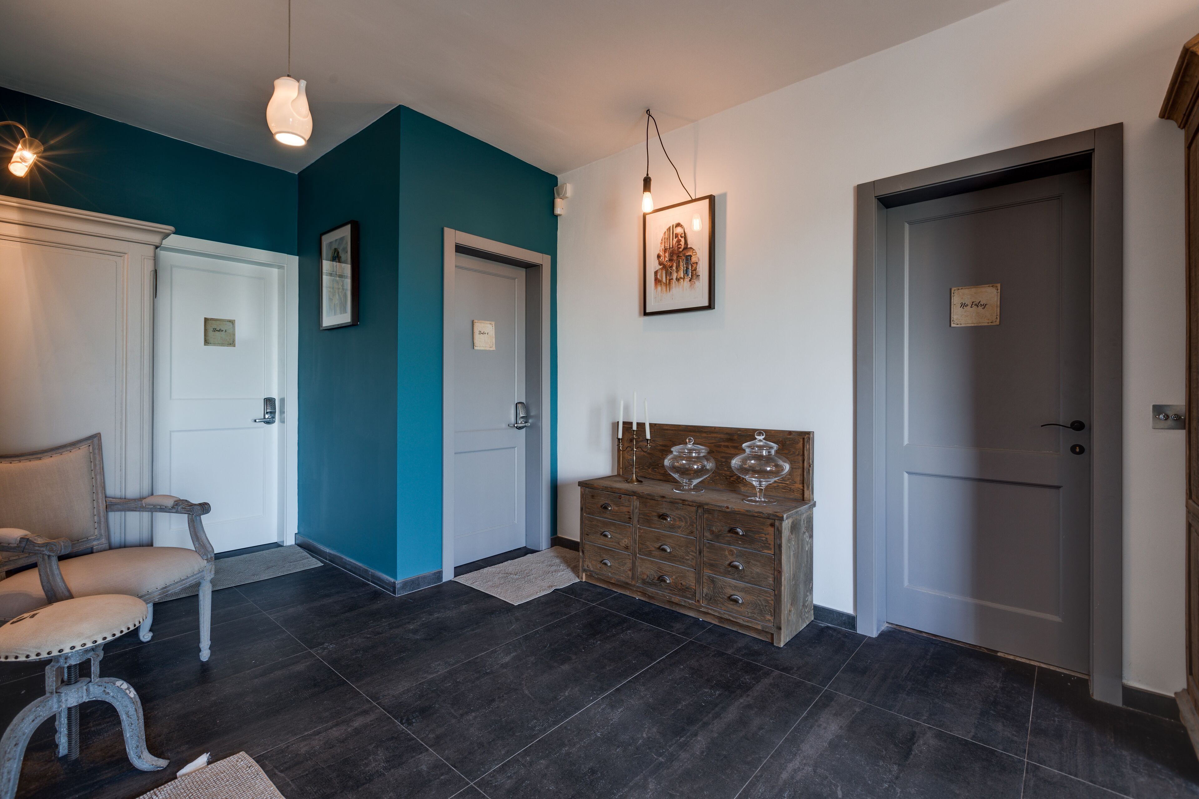 A hallway with a teal accent wall, a wooden bench, a white door, and a wooden dresser with glass containers.