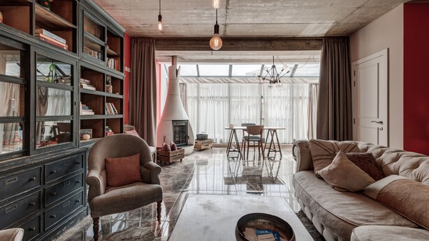 Amplia sala de estar con un sofá, un sillón y una mesa de centro. Se observa una estantería llena de libros y objetos decorativos. Al fondo, un hogar a leña, y una ventana grande con cortinas.