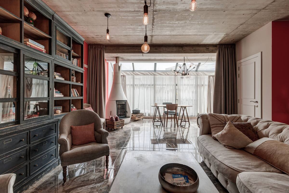 Ein geräumiges Wohnzimmer mit Sofa, Ohrensessel und Couchtisch. Ein Bücherregal ist mit Büchern und Dekoration gefüllt. Im Hintergrund ist ein Kamin zu sehen, und das Zimmer hat ein großes Fenster mit Vorhängen.