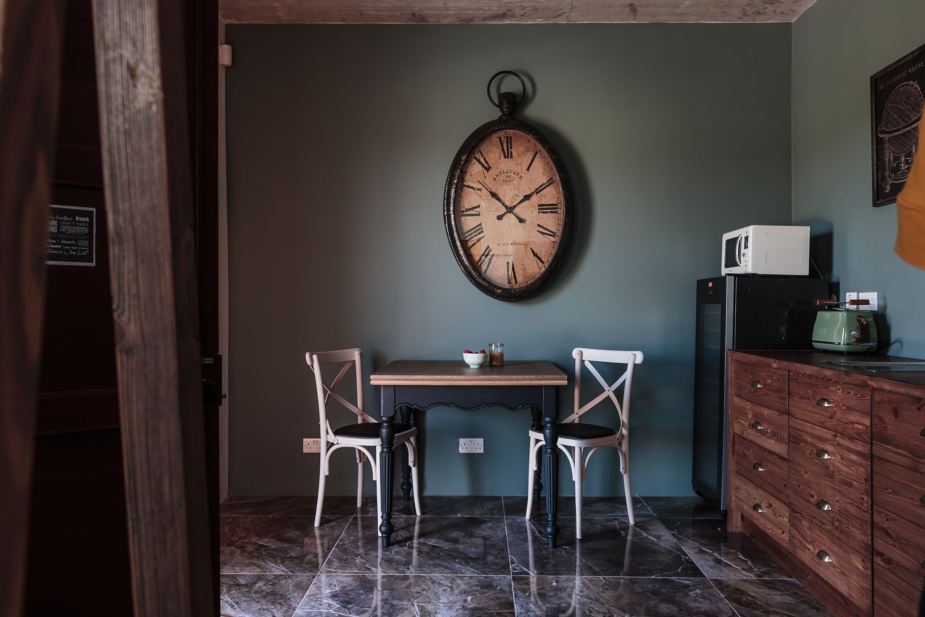 Une pièce avec une table en bois, deux chaises, une horloge au mur, un four à micro-ondes et un meuble.