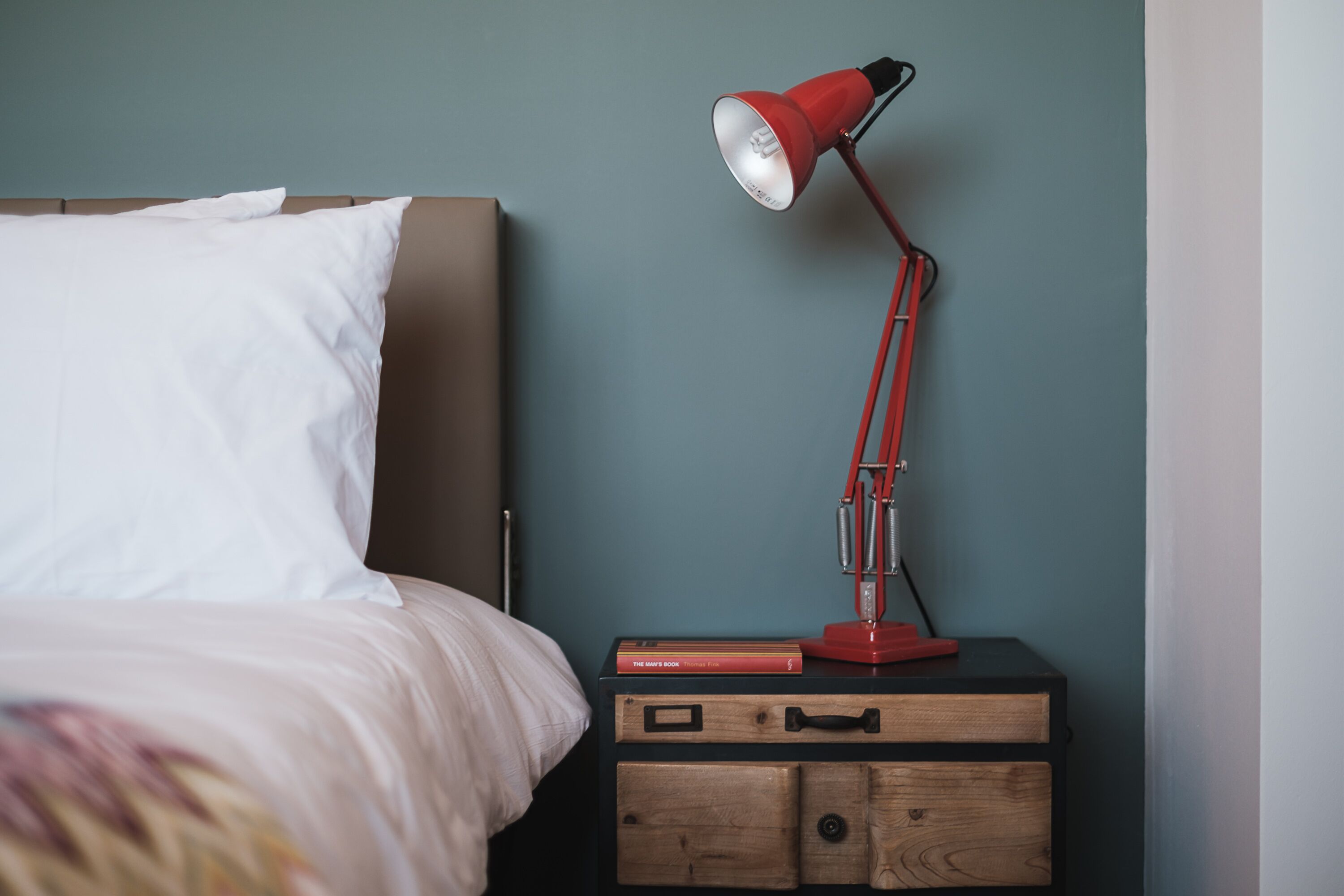 A neatly made bed with a white pillow and a colorful blanket, a red desk lamp, and a book titled "The Game Book" on a wooden nightstand against a teal wall.