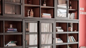 Una habitación con una amplia librería de madera oscura llena de libros y objetos decorativos, dos sillones tapizados y una bombilla colgando del techo.