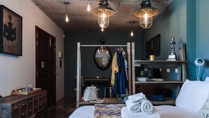 Habitación de hotel con cama, cómoda de madera, espejo, perchero con ropa, estante con objetos y un reloj de pared.