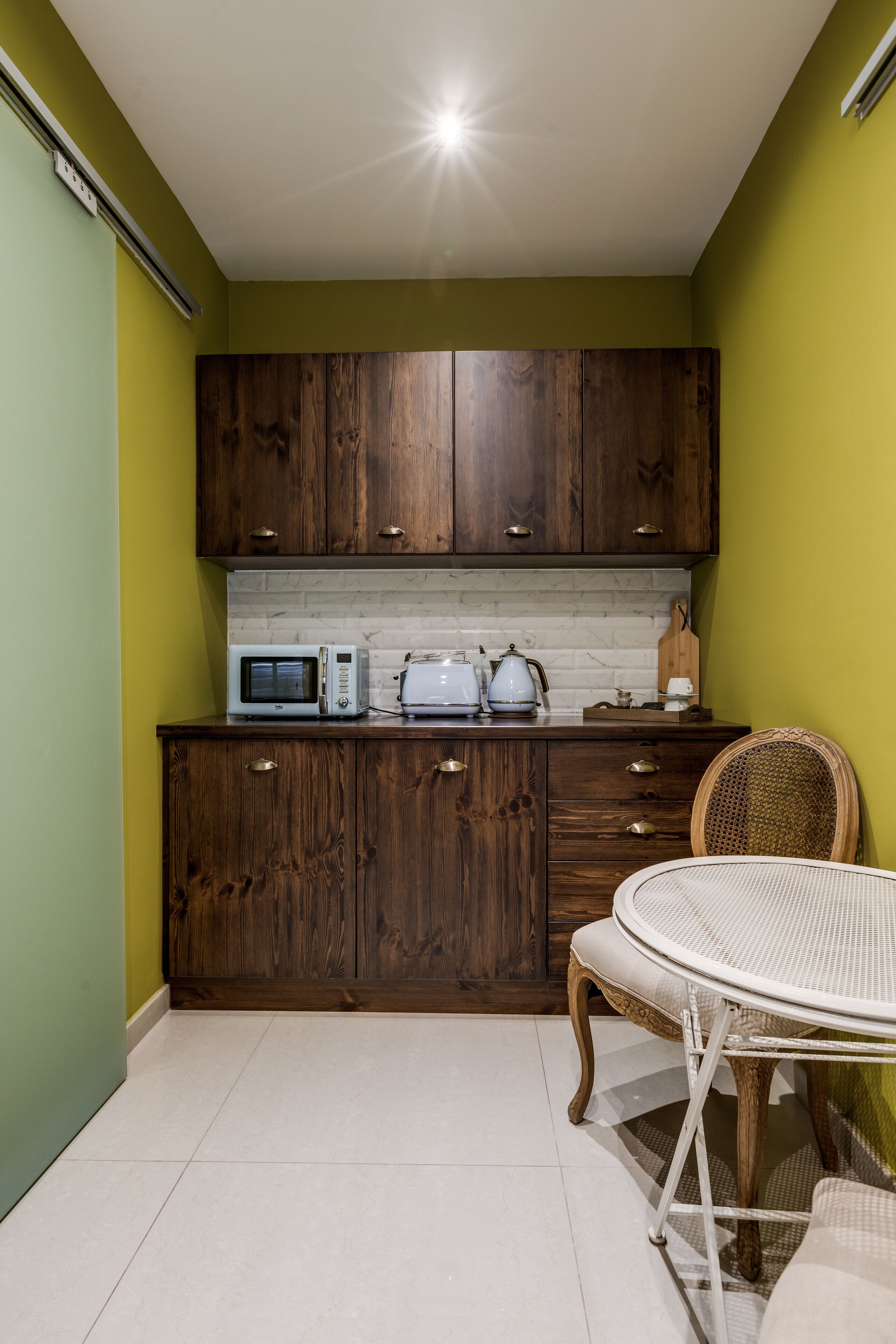 A compact kitchen with wooden cabinets, a microwave, a toaster, and a wicker chair.