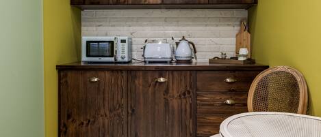 Cocina compacta con armarios de madera, microondas, tostadora y una silla de mimbre.