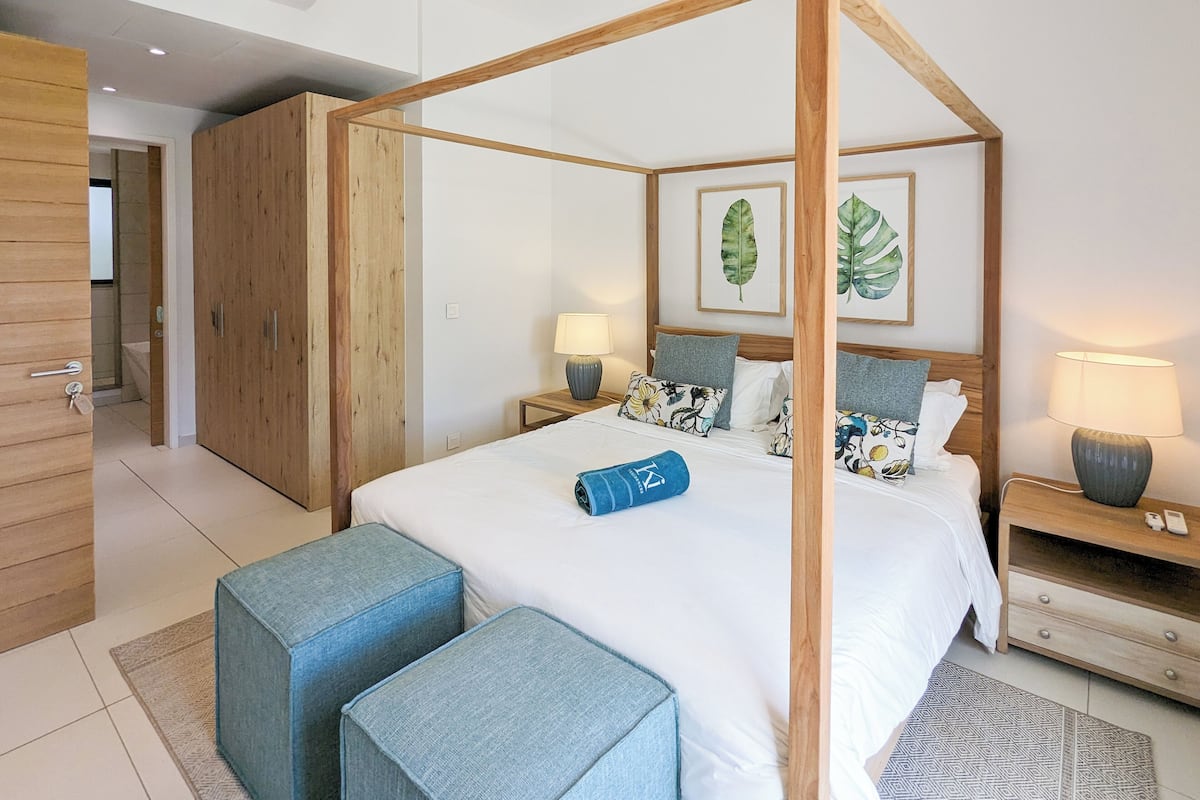 Un dormitorio moderno con una cama con dosel, armario de madera y un baño visible al fondo.