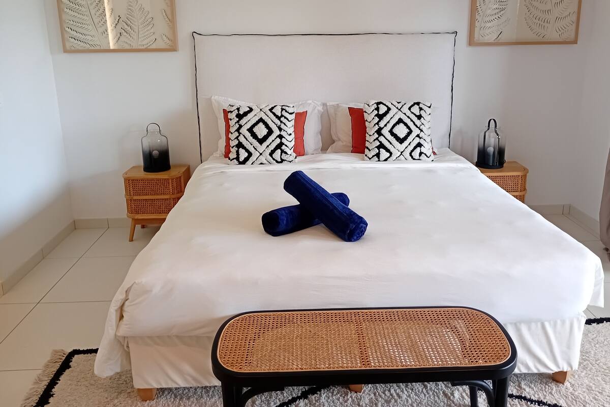 Una cama bien tendida con almohadas estampadas, una mesita de noche con lámpara y una obra de arte colgada en la pared.