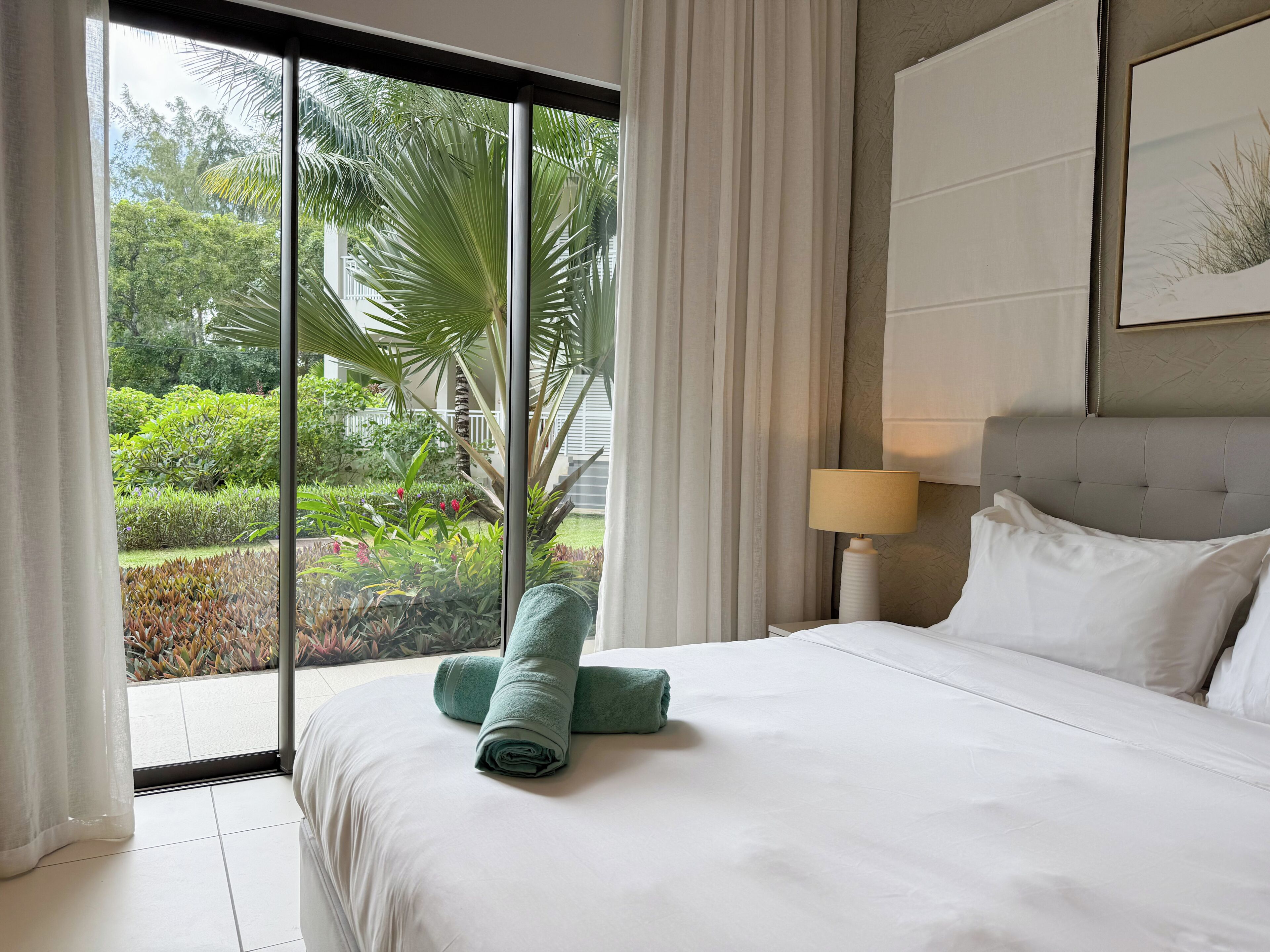 A hotel room with a large window, a bed with white linens, a green towel, a bedside lamp, and a framed picture on the wall.